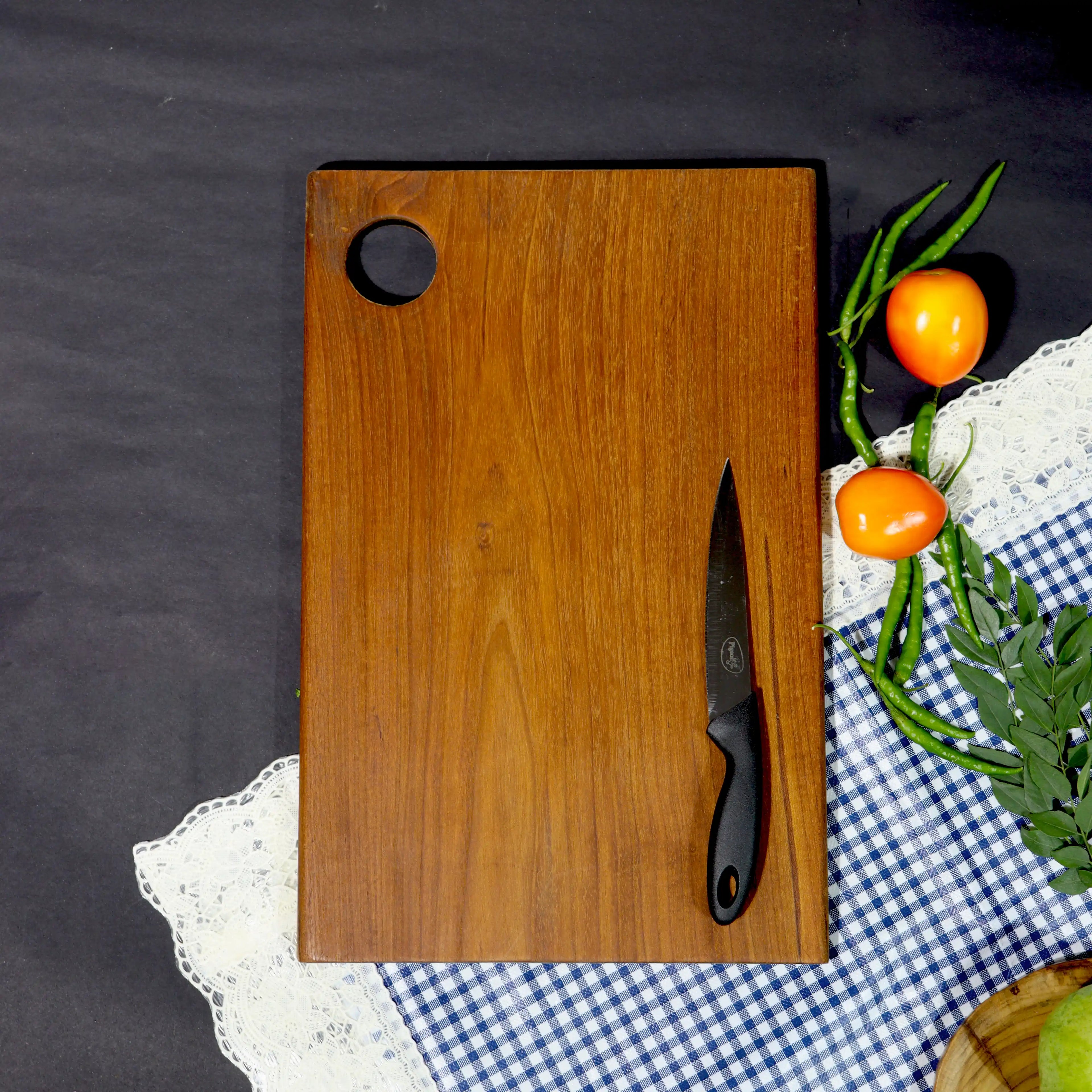 Wooden cutting board with a knife on a checkered tablecloth with vegetables.
